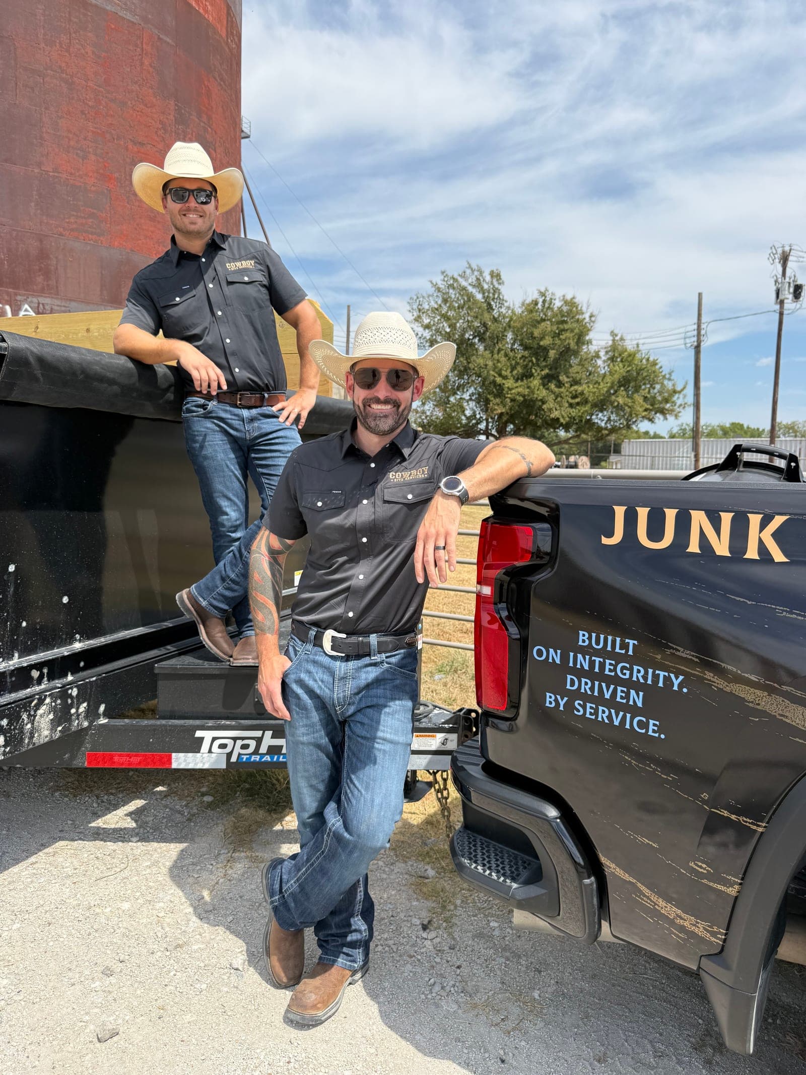 Michael Miller and Daniel Anderson, owners of Cowboy Site Services, in straw cowboy hats beside the company truck and dump trailer